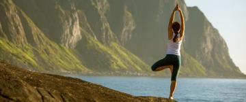 Back of woman standing on one leg in yoga pose in front of a body of water